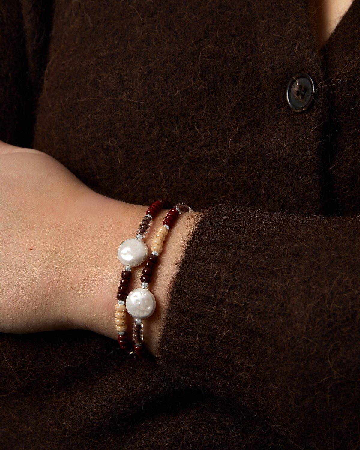 A close-up of a person's wrist wearing two beaded bracelets with white pearls and beige and brown glass beads