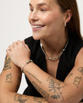 Woman wearing a necklace, bracelet, and earrings with tattoos on her arms against a plain background