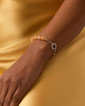 A close-up of a person's wrist wearing a beaded bracelet with peach-orange beads and a silver-toned toggle clasp, set against a yellow satin fabric background.